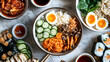 © Nataliia - Assorted Asian cuisine featuring a colorful bibimbap bowl with rice, vegetables, mushrooms, and a soft-boiled egg, surrounded by sushi, ramen, dumplings, kimchi, and dipping sauces on a marble surface