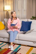 © zinkevych - Positive mature woman in pink sitting on a sofa with a laptop