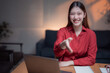 © MINAE - Young Asian businesswoman wearing a vibrant red shirt, shaking hands at her desk in a cozy home office, smiling warmly and engaging with the camera, exuding confidence and professionalism