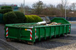 © Viktor - A green dumpster is located in an urban setting, filled with various construction debris and materials. Surrounding foliage is visible, indicating the presence of nearby buildings and infrastructure.