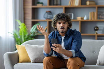 A young man looks confused while holding his smartphone, questioning a message or notification, seated on a couch in a home setting.