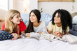 © Xavier Lorenzo - Three cheerful multi-ethnic friends are lying on a bed, smiling and enjoying a slumber party together. Female friendship and youth concept