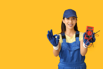 Wall Mural - young female electrician with multimeter showing ok gesture on yellow background