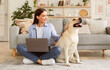 © Prostock-studio - Everyday Life Concept. Portrait of smiling woman sitting on the floor and holding pc on lap, having rest with pet. Casual lady stroking her labrador retriever spending time in living room at home