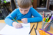 © Niko_Dali - A young boy in a blue shirt sits outdoors, drawing with colored pencils. The child is focused on landscape art. The wooden table has charcoal, pencils, ruler, and scissors.