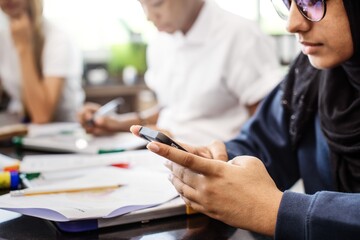 Fototapete - Asian female student in hijab using smartphone in class. Student focused on smartphone. Other students in background. Classroom setting with students and smartphones. Muslim teen student in classroom.