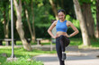 © crizzystudio - Happy fitness woman warming up her legs before running, enjoying a refreshing morning workout in a vibrant green park filled with trees and benches in the background