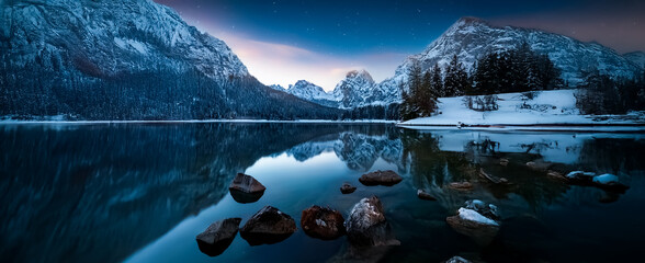 Aufkleber - Snowy mountains reflect in a tranquil lake beneath a twilight sky. Rocks dot the foreground in this peaceful winter landscape scene.
