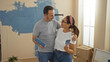 © Krakenimages.com - Middle-aged hispanic couple embracing joyfully in a newly painted living room of their home, holding paint supplies, embodying love and teamwork in indoor home renovation.