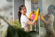 © PaeGAG - Happy asian cleaning lady wearing apron and gloves, holding spray bottle and cleaning glass window with microfiber cloth