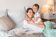 © InsideCreativeHouse - Little cute small boy child son hugging embracing his young mom on the bed while she is reading a book of fairytale for him. Family moments, spending time together with children