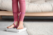 © Pixel-Shot - Young woman with measuring tape around her legs standing on scales at home. Diet concept