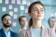 © Platoo studio - confident businesswoman in glasses stands in focus, with blurred team and sticky notes in background