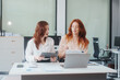 © NanSan - Two Caucasian women work in an office, happily using laptops and computers at a desk, discussing financial projects, analyzing financial statements,collaborating in a meeting to ensure effective