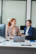 © NanSan - Two Caucasian women work in an office, happily using laptops and computers at a desk, discussing financial projects, analyzing financial statements,collaborating in a meeting to ensure effective