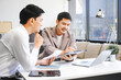 © R Photography - Two Asian businessmen sit in the office using laptops. They manage business tasks, communicate with clients, analyze data, and oversee projects to ensure success and efficiency in their operations