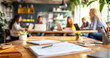 © David Giraud - Des documents sur une table avec un stylo et un cahier, des personnes floues en arrière-plan lors d'une réunion de travail au bureau.