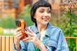 © Iryna - Cheerful woman holding a smartphone while enjoying a sunny day in a park, surrounded by greenery and modern architecture