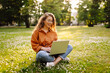 © maxbelchenko - Happy woman with laptop working in a sunny park on a green lawn. Young female freelancer enjoying the weather outdoors. Remote work concept. Active lifestyle.