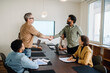 © Vadim Pastuh - Two businessmen shake hands at the end of a successful business meeting. A diverse group of colleagues watches the interaction in a modern conference room.
