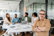 © Vadim Pastuh - Business leader in smart casual attire, sitting confidently at a modern office desk while his team discusses strategies in the background.