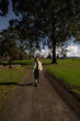 © asier - Young woman walking paved path through verdant park, surrounded by lush greenery and towering trees, basking in sunlight under azure sky with wispy clouds