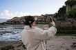 © asier - Female photographer capturing scenic combarro coastal town views using smartphone, framing galician landscape under bright blue sky with scattered clouds