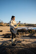 © asier - Female musician performing acoustic guitar, wearing cowboy boots and white fur coat, standing on rocky shoreline during sunset with serene, artistic mood