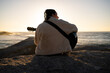 © asier - Female musician playing acoustic guitar, sitting on coastal rock during sunset, overlooking serene ocean waves with peaceful expression