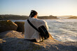 © asier - Young musician with braids sitting on rocky shoreline, embracing acoustic guitar during golden sunset, ocean waves illuminating coastal landscape at punta faxilda