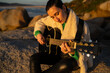 © asier - Female guitarist performing acoustic melody, seated on rocky coastline near punta faxilda, silhouetted against golden sunset lighting coastal landscape
