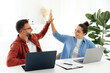 © Kateryna - Cheerful a man and a woman high five while sitting at their desk in a modern office, celebrating a success together, both are working on their laptops and enjoying their time at work, smile