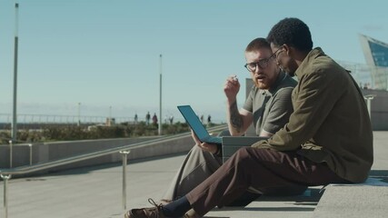 bearded man in short-sleeved shirt and curly-haired African American man in jacket discussing work on laptop while sitting on concrete steps outdoors