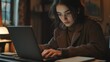 © Denis - The image captures a woman engrossed in her work on a laptop. She is seated comfortably, focused on the screen in front of her. The room around her is dimly lit, suggesting it's late at night. Her