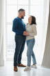 © ninelutsk - Loving couple at 40s stands by large window in home, enjoying quality time together. Family smiles, hug, moment of joy and togetherness