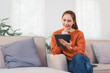 © PRIME STOCK LAB - Asian Businesswoman in Orange Sweater Sits on Sofa, Smiling While Using Tablet in Bright Living Room.