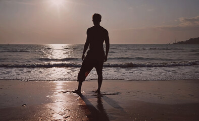 Poster - Man stands on the beach at sunset, looking out at the ocean. The scene is peaceful and serene, with the man's silhouette against the backdrop of the sun and the water