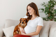 © Inga Bulgakova - A smiling pregnant woman wearing a white shirt sits on a beige couch, gently cradling a small dachshund dog in her arms, with a green plant in the background.