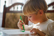© Valua Vitaly - Small kid drawing a house on a table. Young caucasian boy sketching with markers at home on a sunny day. Blonde child coloring a sketch of a house with markers as his homework assignment.
