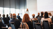 © kasto - Business and entrepreneurship symposium. Female speaker giving a talk at business meeting. Audience in conference hall. Rear view of unrecognized participant in audience.