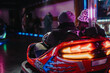 © qunica.com - A couple enjoying a ride together in a red bumper car surrounded by vibrant colorful neon lights and blurred background, evoking feelings of fun, joy, and excitement at an indoor amusement park event.