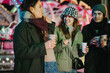 © qunica.com - A group of friends smiling and enjoying hot drinks at a festive outdoor event in winter. Bright lights and cheerful atmosphere create a perfect holiday setting surrounded by joy and warmth.