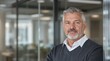 © Damir - Close-up of a confident mature man with gray hair, wearing a sweater and shirt, standing in a modern office environment. Business confidence concept. Ai generative