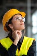 © Iryna - A woman wearing a yellow vest and a hard hat is looking up at something. She is wearing safety glasses and a reflective vest