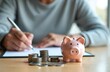 © Vadym - Woman writes on paper near piggy bank, stack of coins. Financial planning, smart savings strategies. Navigating retirement, expert guidance. Wealth, income, confidence. Budgeting, flourishing,