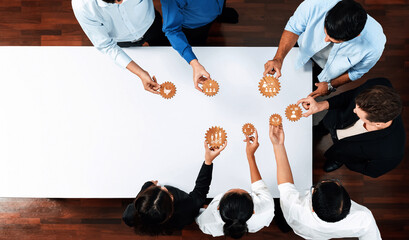 Poster - A diverse group of professionals collaborates around a table, exchanging ideas and holding gears to symbolize teamwork and innovative problem-solving in a business environment. Amity