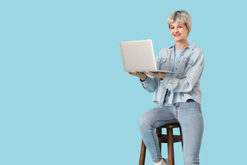 Wall Mural - portrait of beautiful young woman with laptop sitting on chair against blue background