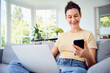 © baranq - Happy woman sitting on sofa at home using smartphone and laptop