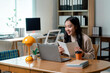© PaeGAG - Young Asian businesswoman attending a video conference holding a notebook and smiling in her home office
