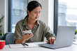 © MIND AND I - Businesswoman sitting at desk on couch in workplace or at home working on laptop and analyzing data on charts and graphs and writing on papers to make business plan and strategies for company,